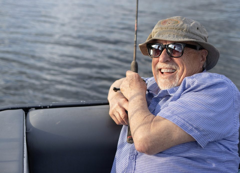 Clyde W. Snyder in a bucket hat and sunglasses, laughing as he braces a bent fishing rod on a boat, with the lake behind him.